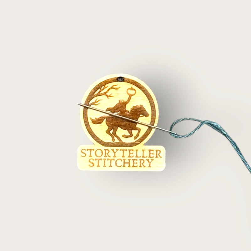 A wood needle minder with an engraved silhouette of a rider on a horse, the Headless Horseman, and the words 'STORYTELLER STITCHERY', with a needle and long teal thread on a plain light background.
