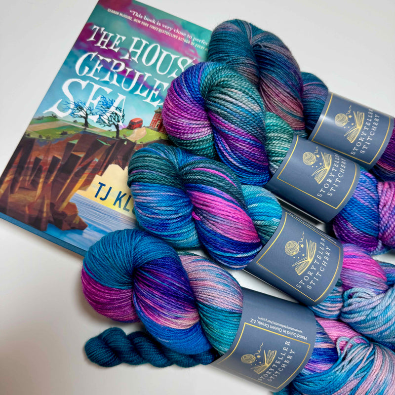Hand-dyed yarn in deep jewel tones with luminous depth, inspired by The House in the Cerulean Sea by T.J. Klune, shown on bases with the book in the background.