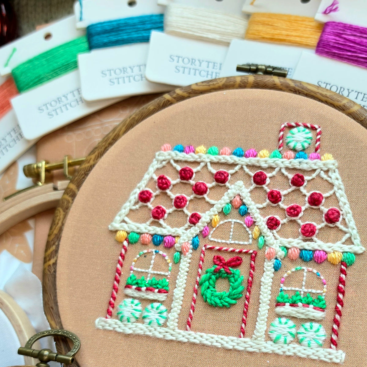 Close-up of a finished gingerbread house embroidery hoop pattern, displayed with colorful hand-dyed stitching thread cards.