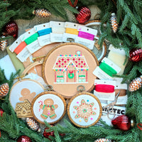 Gingerbread embroidery collection featuring stitched house, man, and star hoops, matching thread cards, and accessories displayed in a festive pine wreath with ornaments.