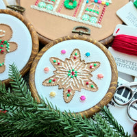 A hand-stitched sugar cookie embroidery pattern in a wooden hoop, alongside gingerbread man and gingerbread house designs, displayed with vibrant hand-dyed thread cards and festive pine needles.