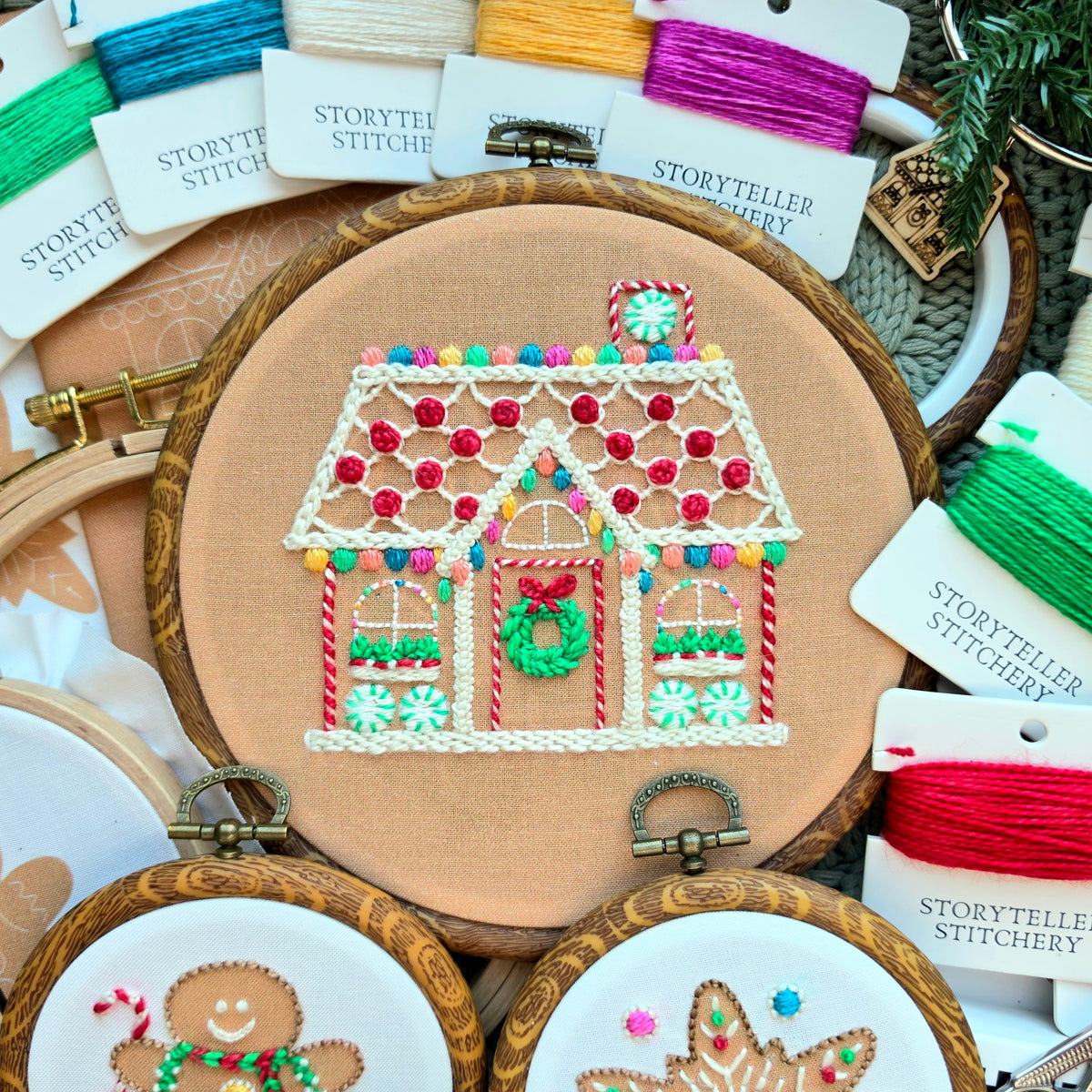 A hand-stitched gingerbread house pattern in a wooden hoop, alongside gingerbread man and star designs, displayed with vibrant hand-dyed thread cards and festive pine needles.