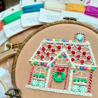 Close-up of a finished gingerbread house embroidery hoop pattern, displayed with colorful hand-dyed stitching thread cards.