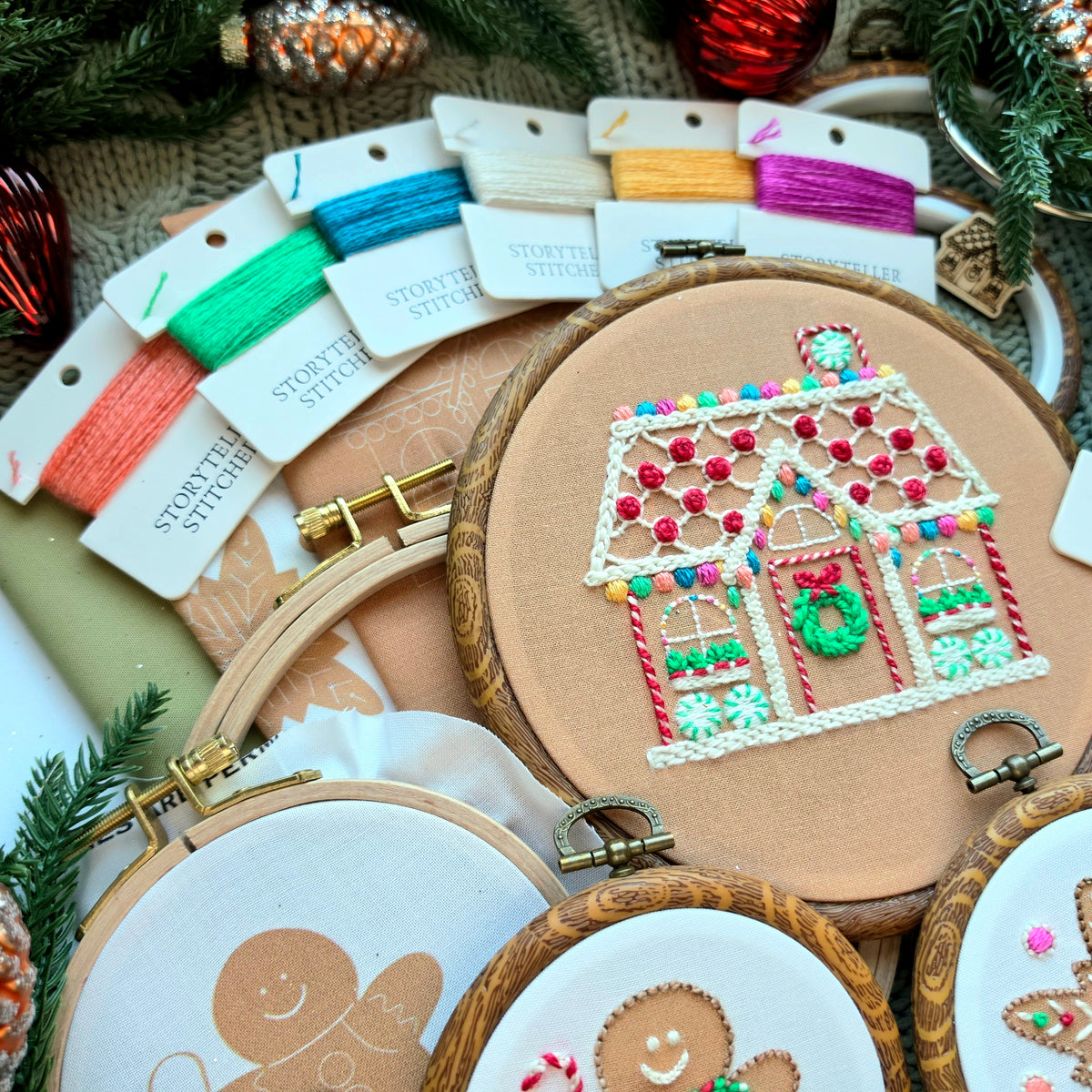 A styled flat-lay of embroidery supplies, featuring wooden embroidery hoops with finished gingerbread house and gingerbread man designs, alongside multiple colorful thread spools from Storyteller Stitchery.