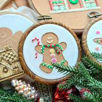 A hand-stitched gingerbread man embroidery pattern in a wooden hoop, alongside gingerbread cookie and gingerbread house designs, displayed with coordinating needle minder and festive pine needles.