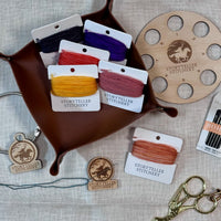 Storyteller Stitchery hand-dyed threads and tools on a textured linen background. The set includes multiple bobbins of threads for needlepoint and embroidery in various colors (black, purple, orange, yellow, copper brown, variegated orange), and various wooden. tools with The Headless Horseman design engraved, including 8 hole thread organizer, floss drop charm, needle and minder. 