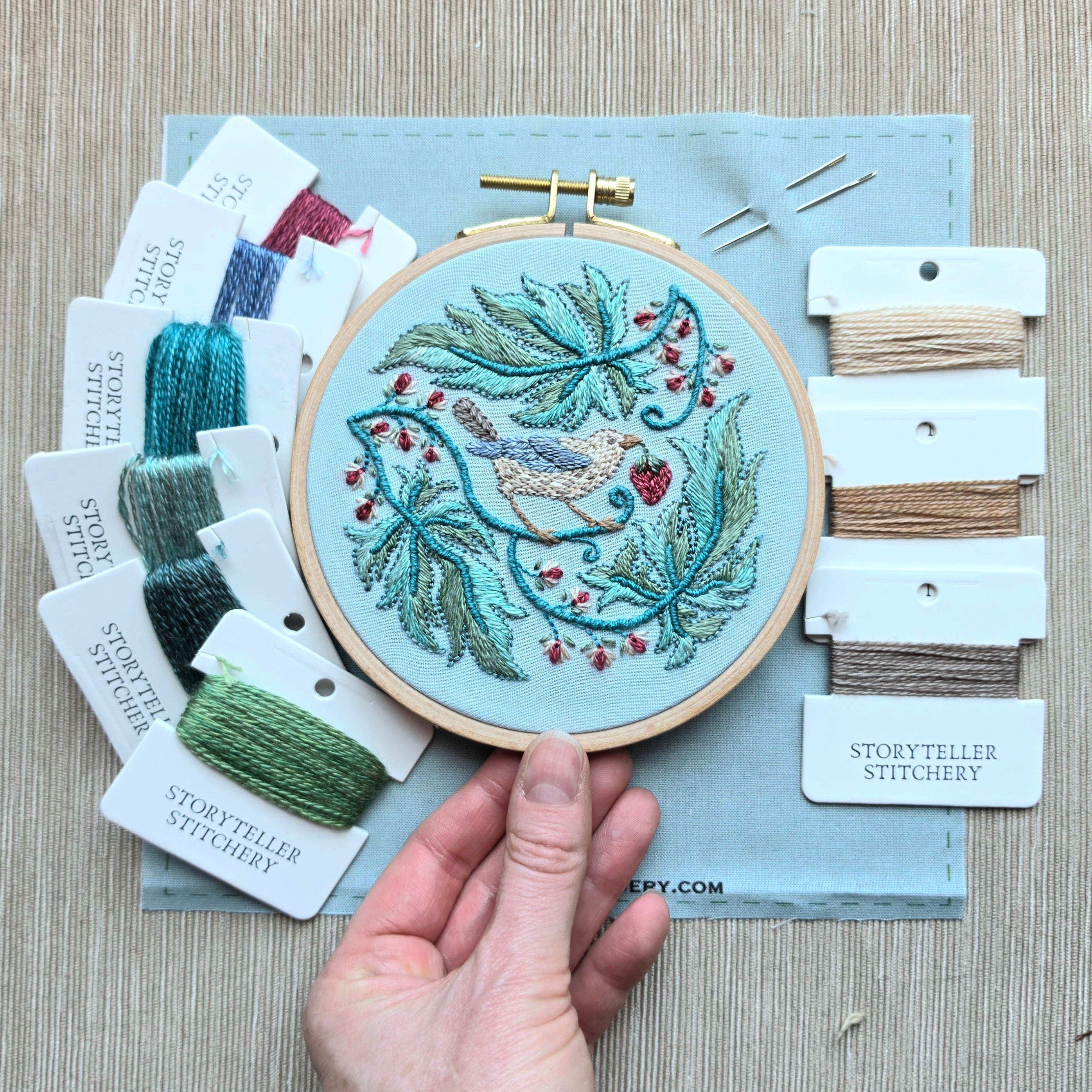 Embroidery hoop with bird design held by a hand, surrounded by embroidery threads and tools.