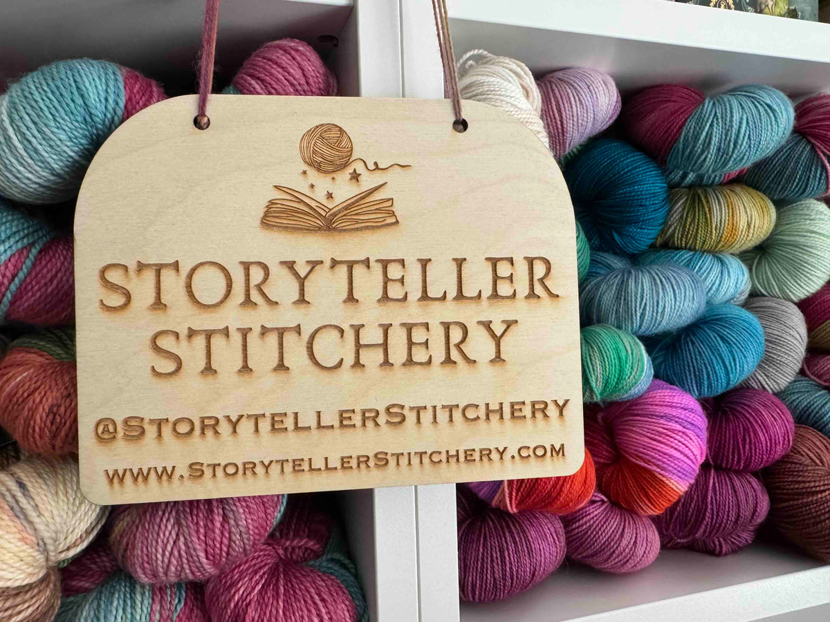 Wooden sign for Storyteller Stitchery surrounded by colorful yarn.